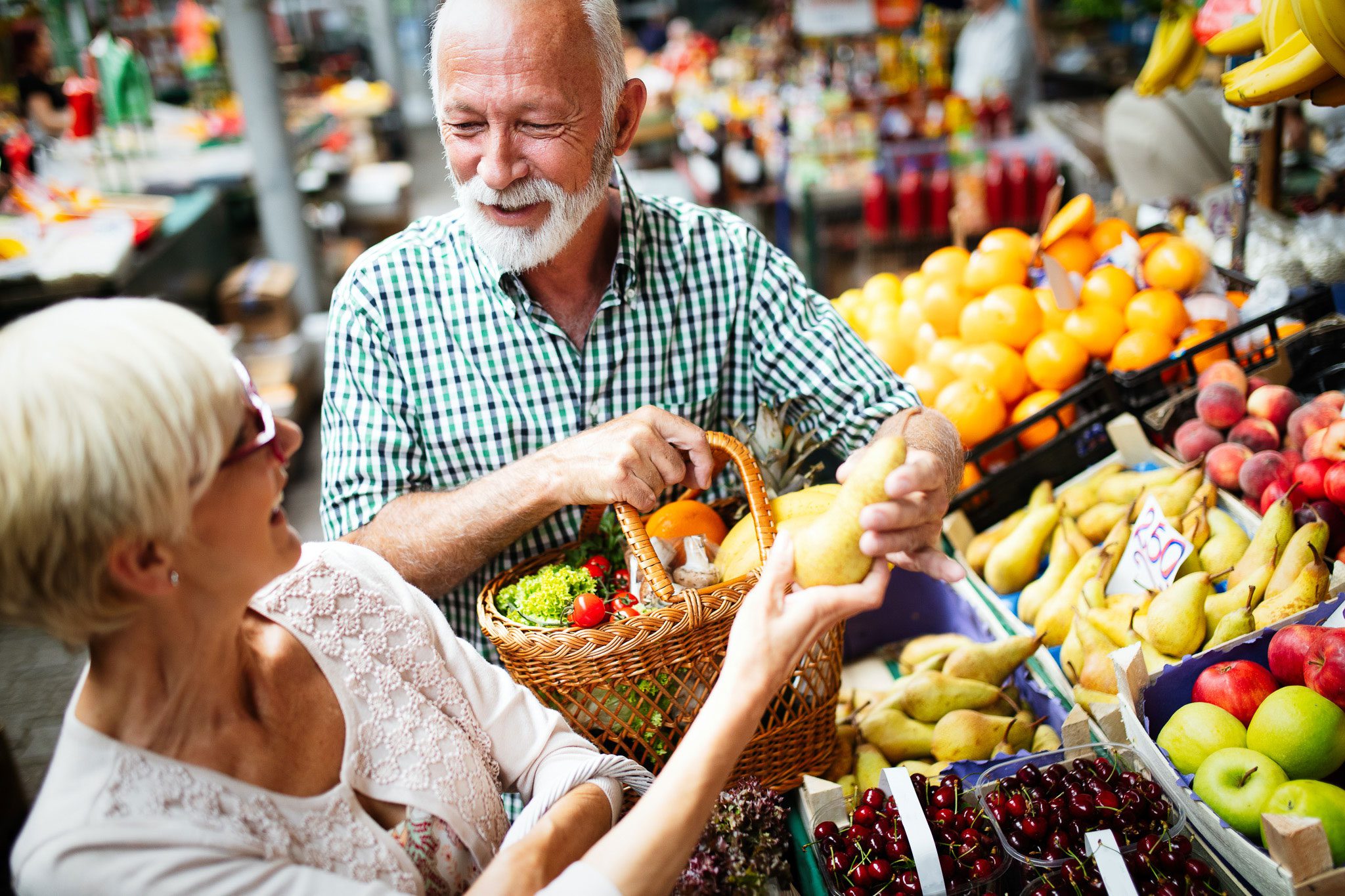 An older couple shops for fresh fruit and vegetables at an outdoor market, smiling as they examine produce and fill a wicker basket.
