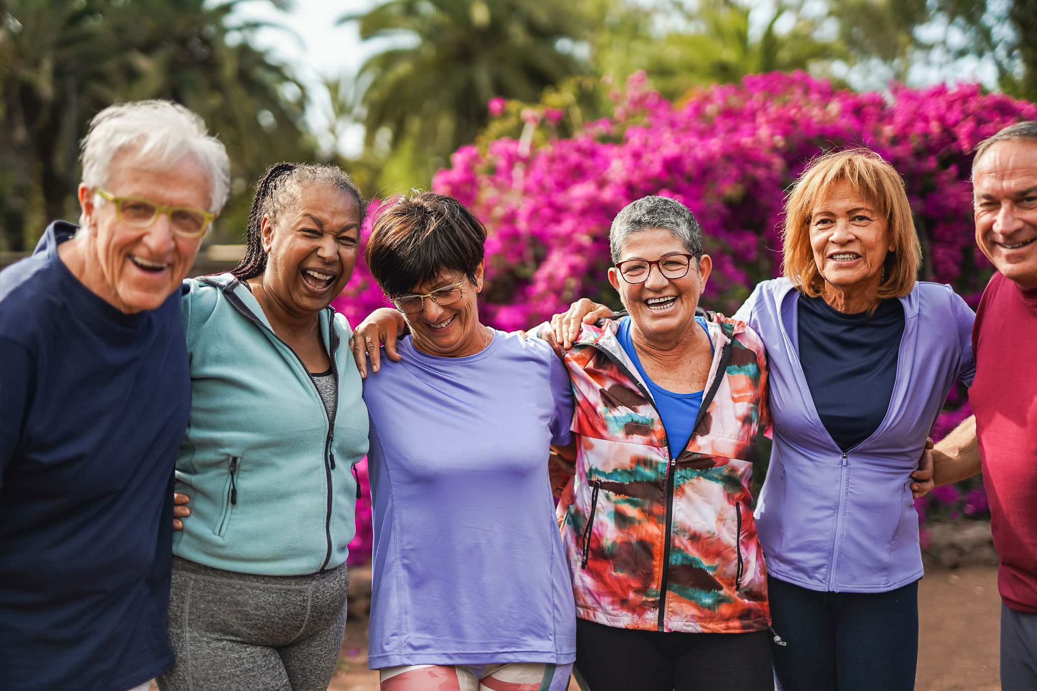 Six older adults stand outdoors in front of blooming pink flowers, smiling and with arms around each other, dressed in casual activewear.